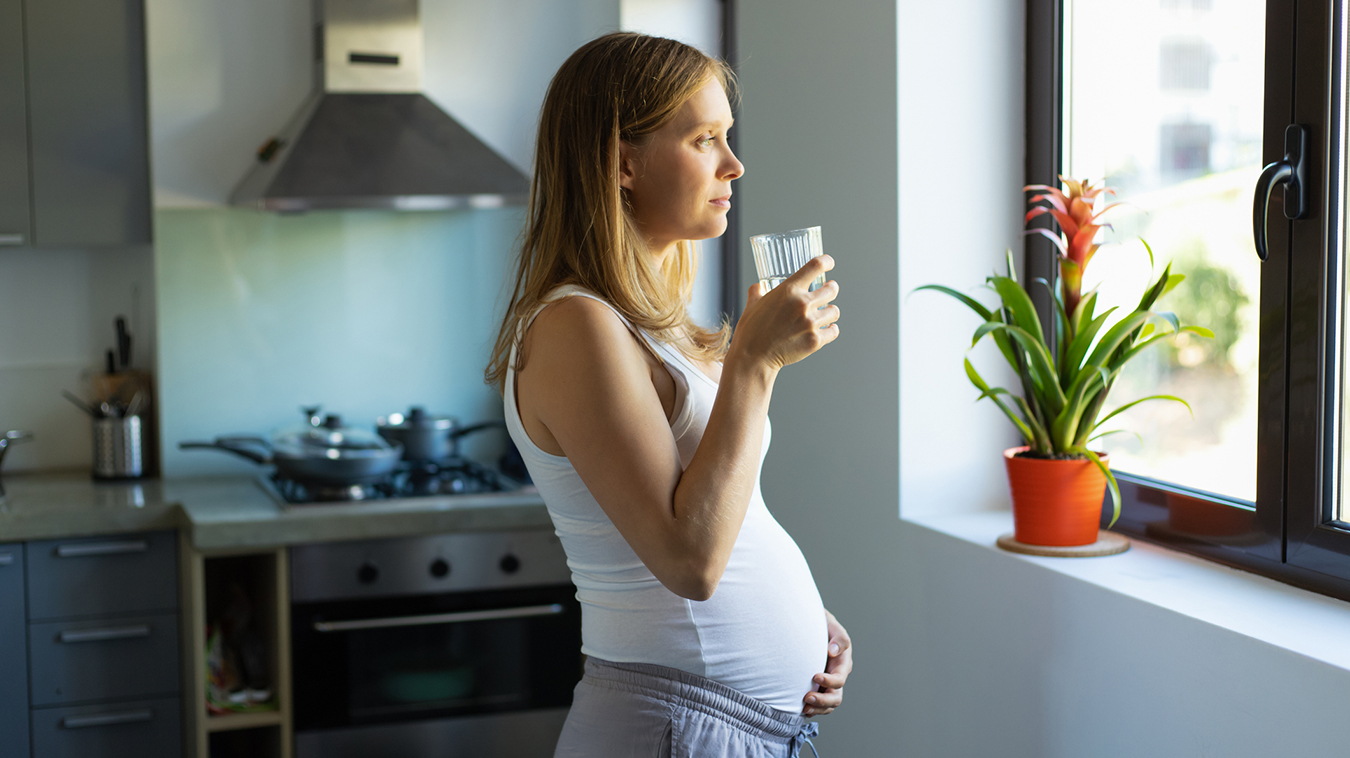 Pensive pregnant woman stares out the window holding a glass of water